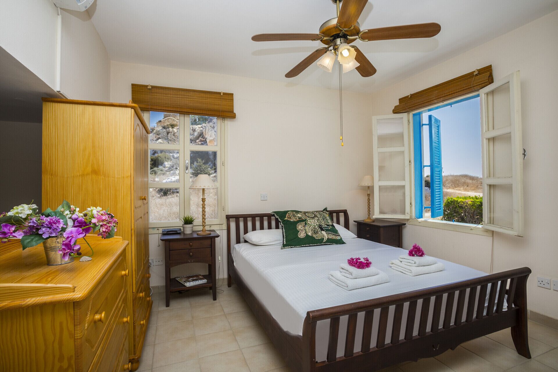 Un dormitorio con cama, mesitas de noche, cómoda de madera, ventilador de techo y ventanas con contraventanas.