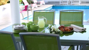 Mesa posta com pratos verdes, copos, jarra de limonada, croissants e flores, com piscina e cadeiras de sol ao fundo.