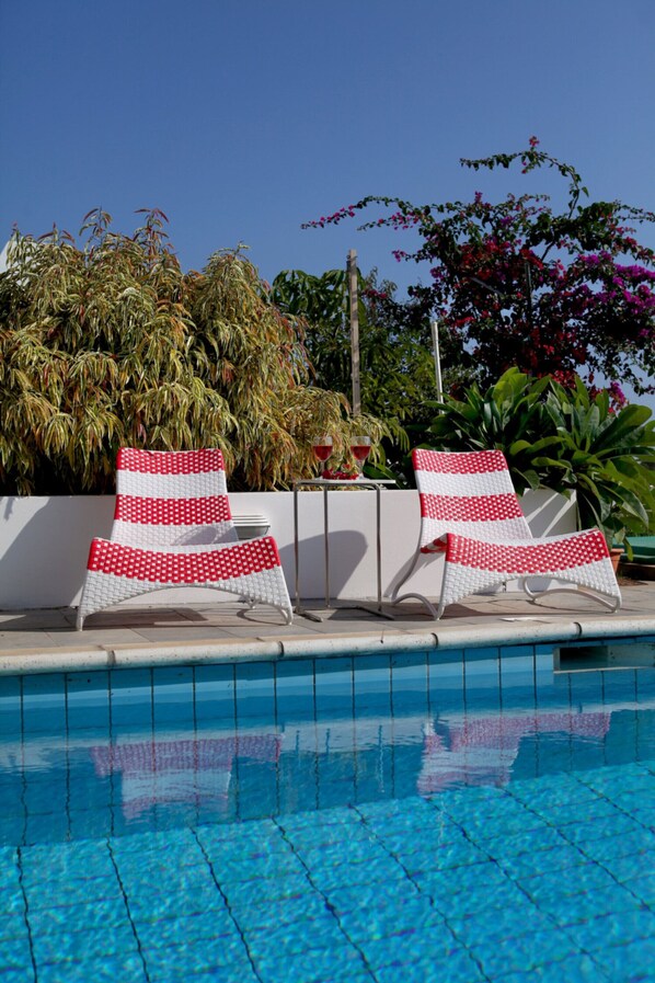 A poolside area with two white lounge chairs with red and white patterns, a white wall, and a variety of plants and trees in the background.