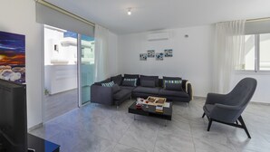 A modern living room with a sectional sofa, a coffee table, and a grey armchair. There is a painting on the wall, a window with curtains, and an air conditioning unit. - At Last You can Rent the Perfect Luxury Villa, Protaras Villa 1494 (Paralimni)