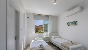 A hotel room with two single beds, a window with a view of the landscape, a bedside table with a lamp, and a wall-mounted air conditioner. - At Last You can Rent the Perfect Luxury Villa, Protaras Villa 1494 (Paralimni)