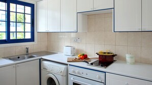 Una cocina con armarios blancos, lavadora, horno y ventana con marcos azules.