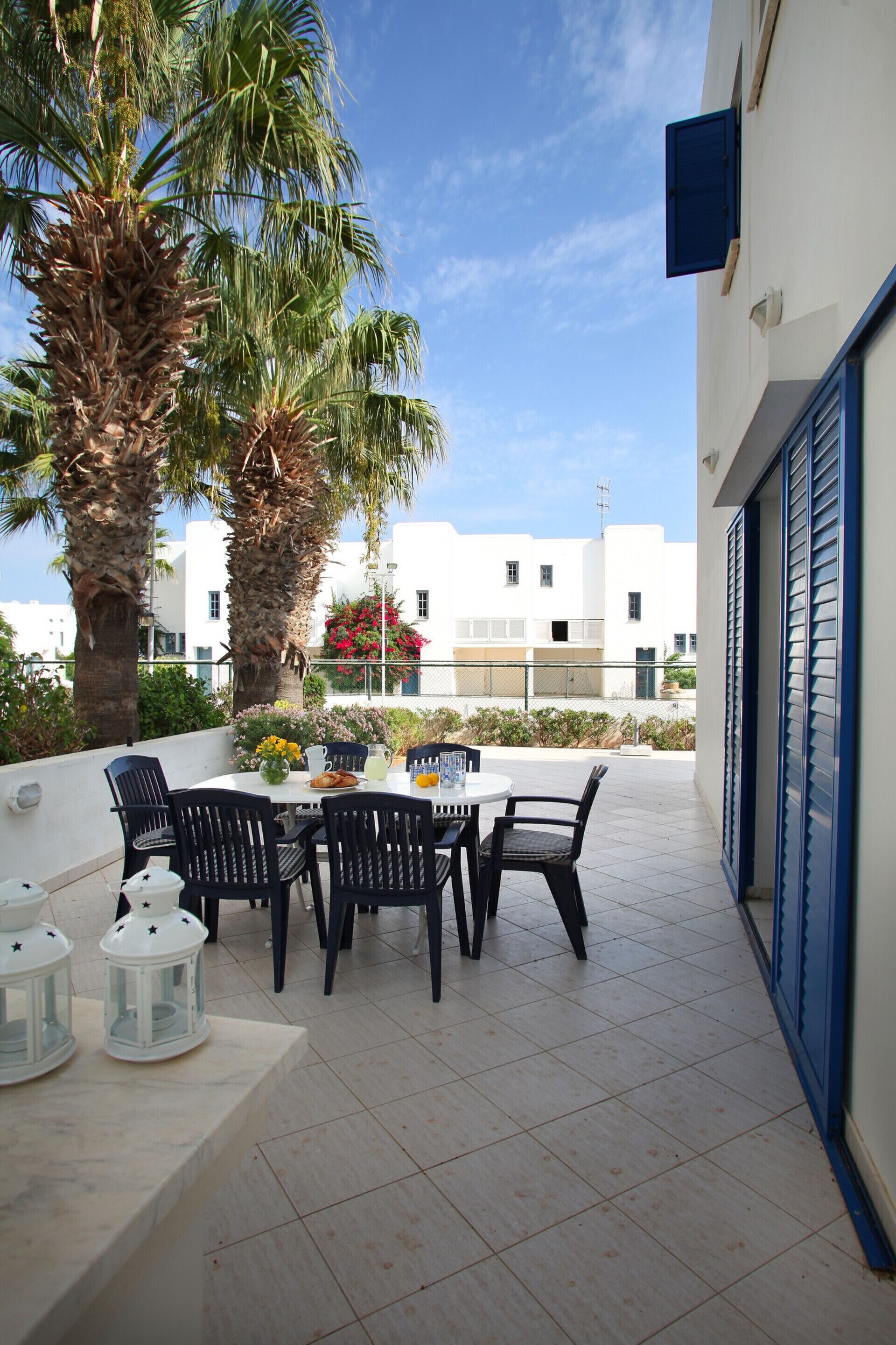 Un patio al aire libre con una mesa y sillas, rodeado de palmeras y un edificio blanco al fondo.