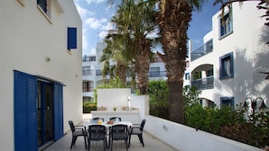 Un patio con una mesa y sillas, rodeado de edificios blancos con balcones, palmeras y suelo de baldosas.