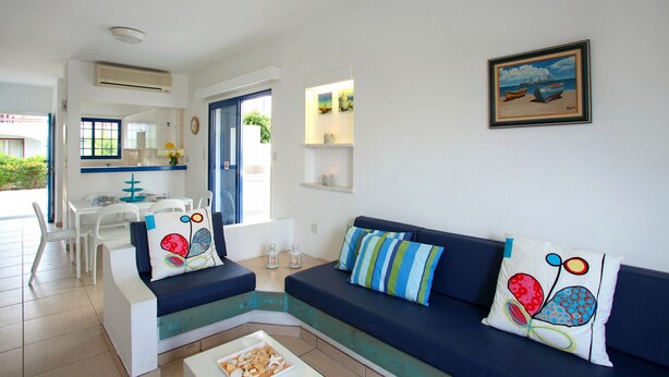 Una sala de estar moderna con un sofá azul, sillones blancos y cojines coloridos. Se observa un comedor con una mesa y sillas blancas, y una cocina visible a través de una puerta abierta. En la pared, cuelga un cuadro enmarcado con una escena de playa.