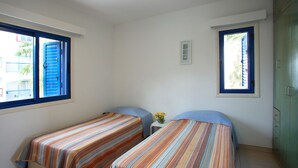 Una habitación pequeña con dos camas individuales, una mesita con un florero y una ventana con contraventanas azules.