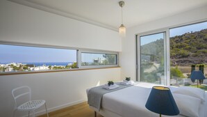 Een slaapkamer met een groot bed, een stoel, een nachtkastje, een lamp en uitzicht op zee en heuvels.