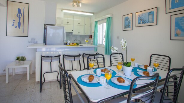 A modern kitchen with white cabinets, a central island, and a dining area with a table set for breakfast. - Indulge in Luxury Living Steps From the Beach,1419 (Paralimni)