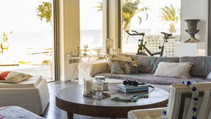 A well-lit living room with a large window overlooking the sea, a round coffee table with books and decorative items, and a comfortable seating area with cushions. - You and Your Family Will Love This Beachfront Villa, Protaras Villa 1526 (Paralimni)