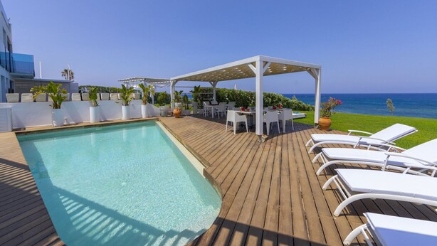 A poolside area with a wooden deck, lounge chairs, and a covered seating area overlooking the sea. - You and Your Family Will Love This Beachfront Villa, Protaras Villa 1526 (Paralimni)