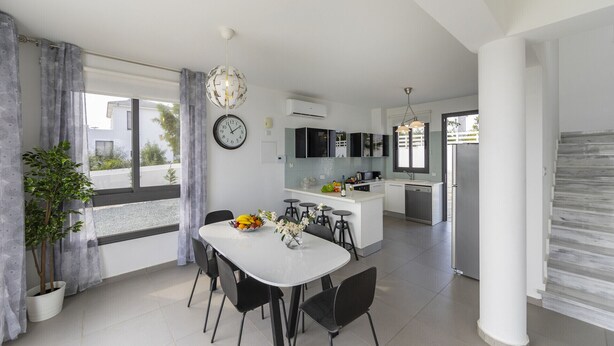 A modern kitchen with white cabinets, a central island, and a dining area with a white table and black chairs. - The Secret to Enjoying Your 5 Star Villa With Large Private Pool, Protaras Villa 1493 (Paralimni)