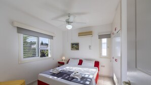 Un dormitorio con cama, ventilador de techo, ventana con persianas, mesita de noche con lámpara y puerta con manija.