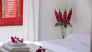 A neatly made bed with white linens and a single pillow, a window with red shutters, and a vase with red flowers on the bedside table. - Discover Perfect Holiday Escape in Protaras, 1411 (Paralimni)