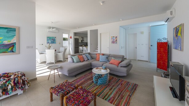 A modern living room with a grey sectional sofa, colorful cushions, a round coffee table, and a vibrant rug. There is a dining area with white furniture and a kitchen in the background. - The Ultimate Guide to Renting Your Luxury 3 Bedroom Villa Minutes From the Beach, Protaras Villa 1530 (Paralimni)
