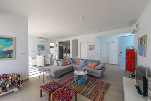 A modern living room with a grey sectional sofa, colorful cushions, a round coffee table, and a vibrant rug. There is a dining area with white furniture and a kitchen in the background. - The Ultimate Guide to Renting Your Luxury 3 Bedroom Villa Minutes From the Beach, Protaras Villa 1530 (Paralimni)