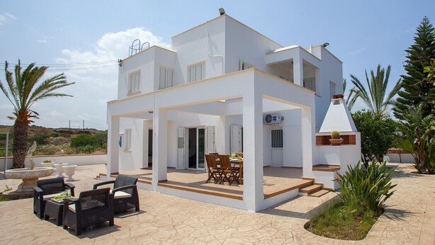 A two-story white building with a covered patio area, outdoor seating, and a small garden. - Imagine Your Family Renting This Luxury Villa With Large Private Pool, Protaras Villa 1543 (Paralimni)