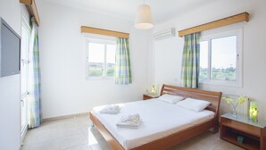 A bedroom with a wooden bed, two windows with curtains, a bedside table with a lamp, and a television on the wall. - Imagine Your Family Renting This Luxury Villa With Large Private Pool, Protaras Villa 1543 (Paralimni)