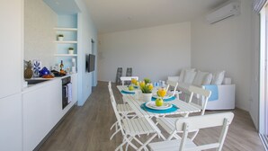 Une cuisine moderne avec des meubles blancs, un coin repas avec des chaises blanches et une table dressée pour deux personnes, et un canapé blanc dans l’espace salon.