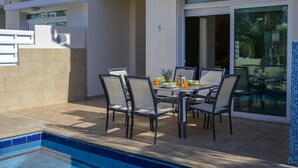 Zona de comedor junto a la piscina con una mesa dispuesta para cuatro personas, rodeada de sillas.