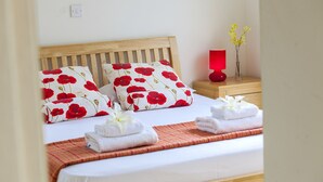 A neatly made bed with white bedding and red floral pillows, a wooden headboard, a bedside table with a red lamp, and a vase with yellow flowers. - Unforgettable 5-star Escape: Protaras Villa 1476 (Paralimni)