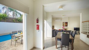 A spacious living area with a dining table and chairs, a TV, and a fireplace. There is a pool visible through an open door. - Unforgettable 5-star Escape: Protaras Villa 1476 (Paralimni)