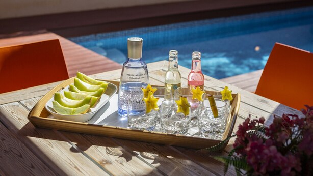 Ein Tablett mit einer Flasche mediterranen Gins, einer Flasche Weißwein, einer Flasche Rosé und einer Melone, platziert auf einem Holztisch am Pool.