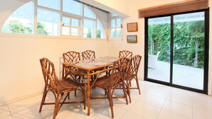 A dining area with a round table and six wicker chairs, large windows with a view of greenery, and a sliding glass door. - The Ultimate Guide to Renting Your Luxury 3 Bedroom Villa Minutes From the Beach, Protaras Villa 1496 (Paralimni)