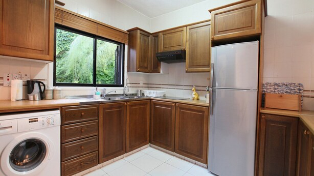 A compact kitchen with wooden cabinets, a stainless steel refrigerator, a built-in washing machine, and a window with blinds. - The Ultimate Guide to Renting Your Luxury 3 Bedroom Villa Minutes From the Beach, Protaras Villa 1496 (Paralimni)