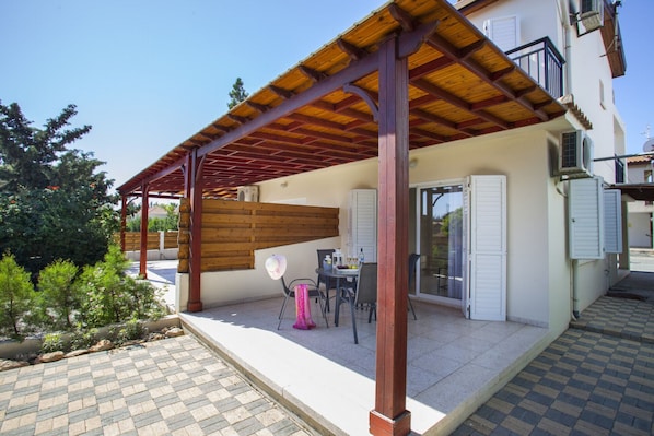 A covered patio area with a table and chairs, a wooden fence, and a view of a building with a balcony. - Stunning Family Villa in Larnaca, 1387 (Pervolia)
