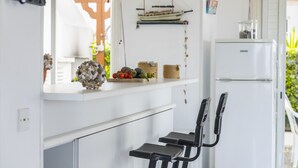 Ett kompakt kök med en vit bänkskiva, en hängande båtdekoration, en liten hylla med frukt och en dekorativ skål samt en svart stol.