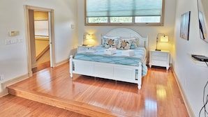A bedroom with a wooden floor, a white bed, two bedside tables with lamps, a window with blinds, and a door leading to another room. - Oceanfront / Sleeps 16 / Beach Days / Screen Porch (Isle of Palms)