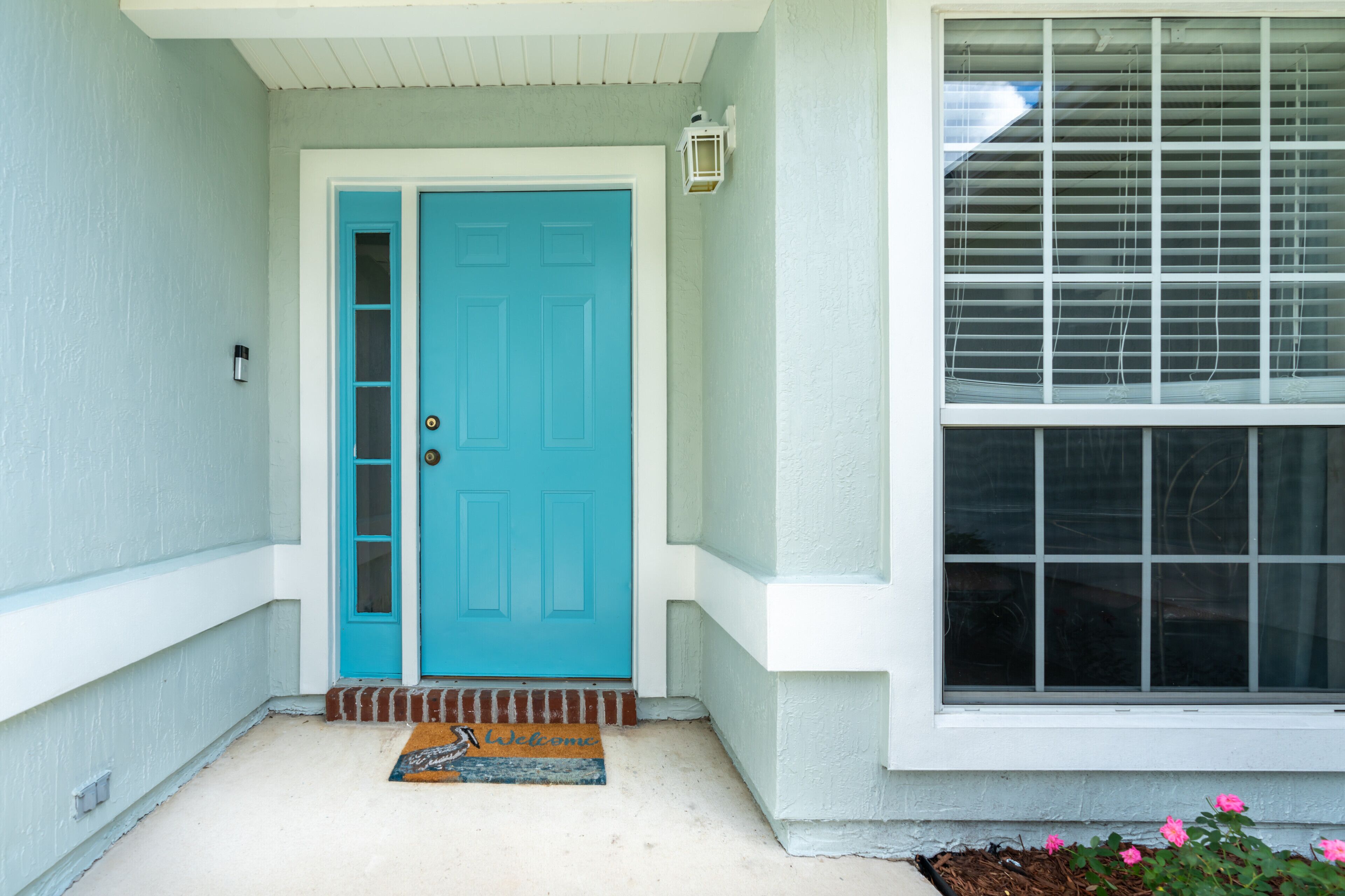 Een turquoise deur met een deurmat, een raam met zonwering en een kleine plantenbak met roze bloemen.