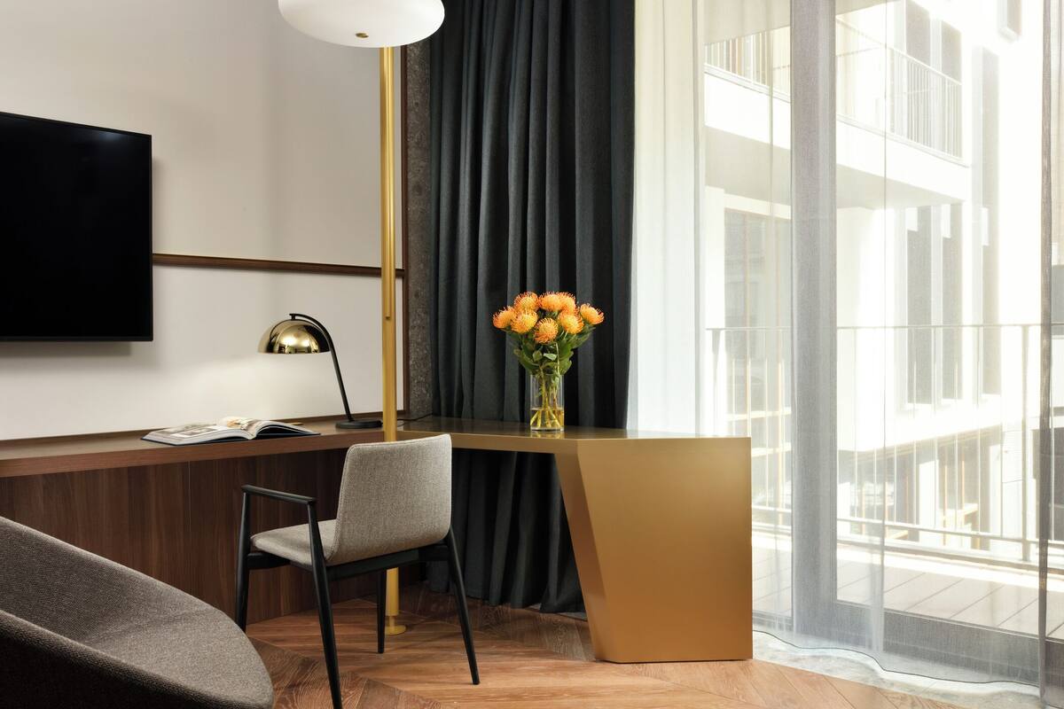 Une chambre d’hôtel moderne avec un bureau en bois, un fauteuil confortable, un téléviseur à écran plat, un vase de fleurs et une grande fenêtre donnant sur la ville.