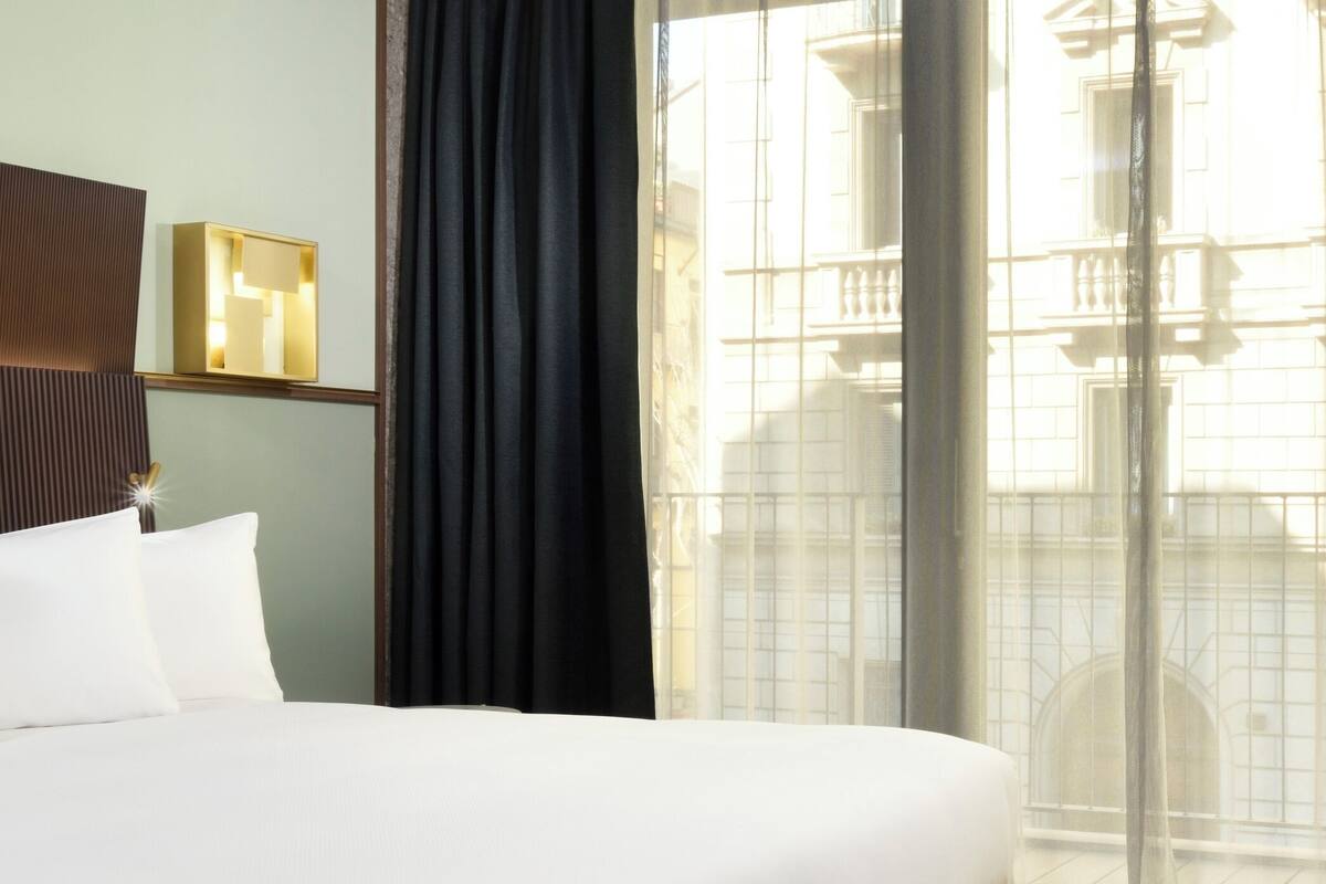 Une chambre d’hôtel avec un grand lit, une table de chevet avec une lampe, une fenêtre donnant sur un immeuble et un rideau.
