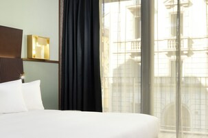 Una habitación de hotel con una cama grande, una mesita de noche con lámpara, una ventana con vista a un edificio y una cortina.
