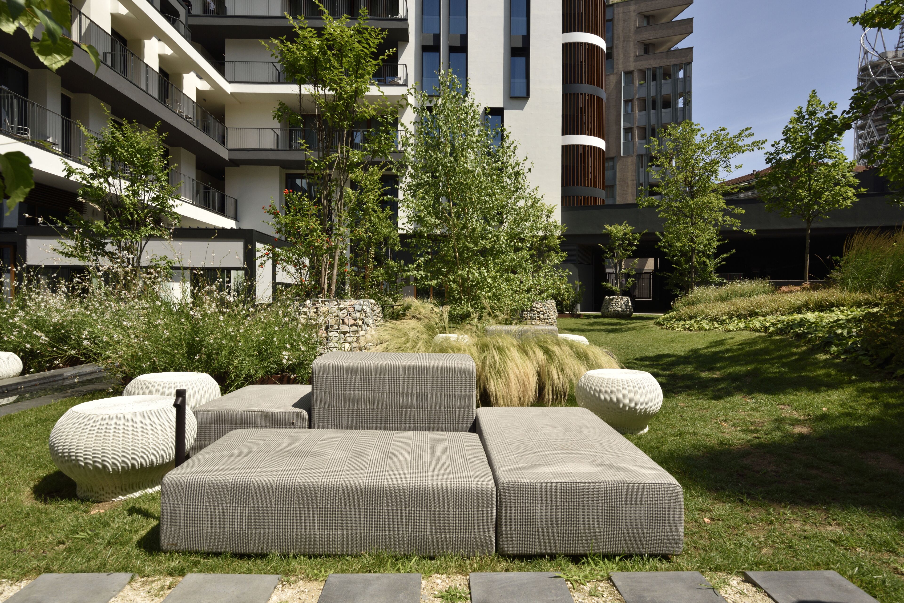 Una moderna zona relax esterna con divani e fioriere, immersa in un giardino curato e affiancata da un edificio a più piani con balconi.