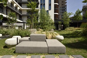 Una moderna zona relax esterna con divani e fioriere, immersa in un giardino curato e affiancata da un edificio a più piani con balconi.