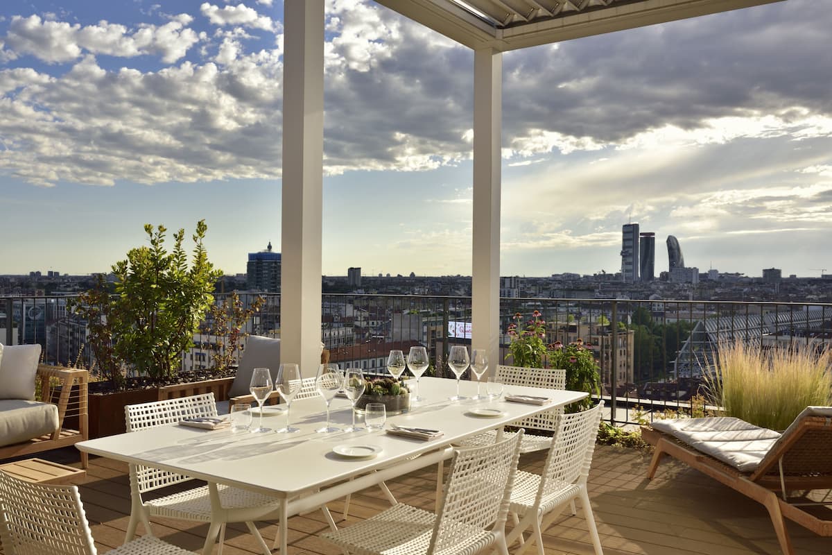 Une terrasse sur le toit, avec une table dressée pour le dîner, entourée de meubles en osier blanc, et offrant une vue sur un paysage urbain avec des bâtiments modernes.