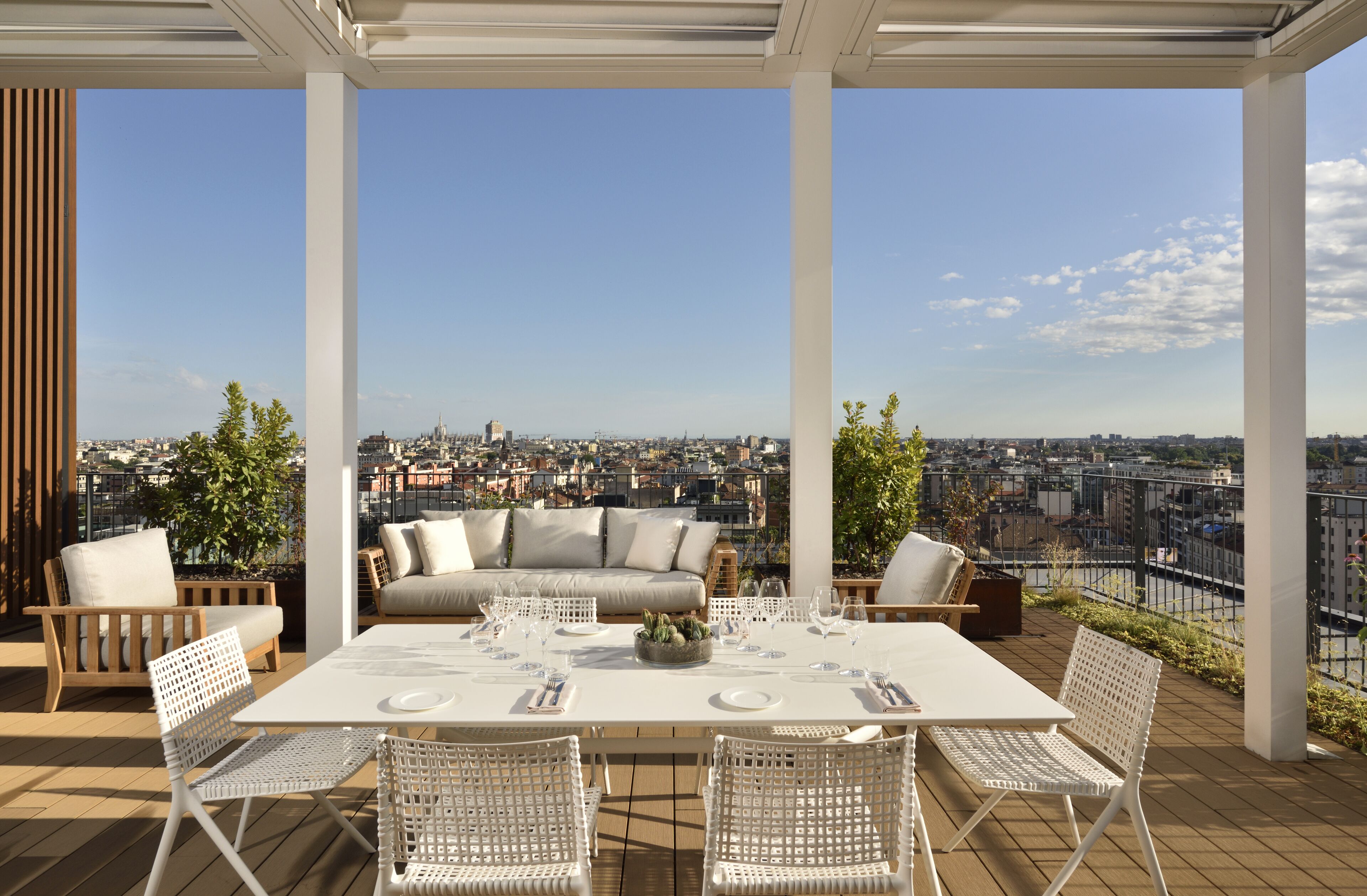 A rooftop terrace with a white table set for dining, surrounded by wicker chairs, and a sofa with cushions.