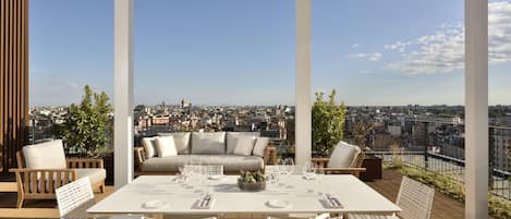 Una terraza en la azotea con una mesa blanca dispuesta para comer, rodeada de sillas de mimbre y un sofá con cojines.