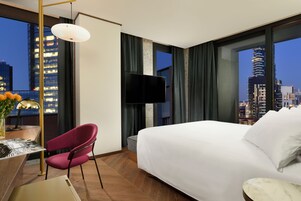 Camera d'albergo con un letto grande, una scrivania con una lampada, una sedia, una televisione e vista sulla città al crepuscolo.