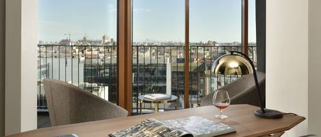 Un espacio de oficina moderno con un amplio ventanal que da a un paisaje urbano, un escritorio con un libro y una copa de vino, y una lámpara.