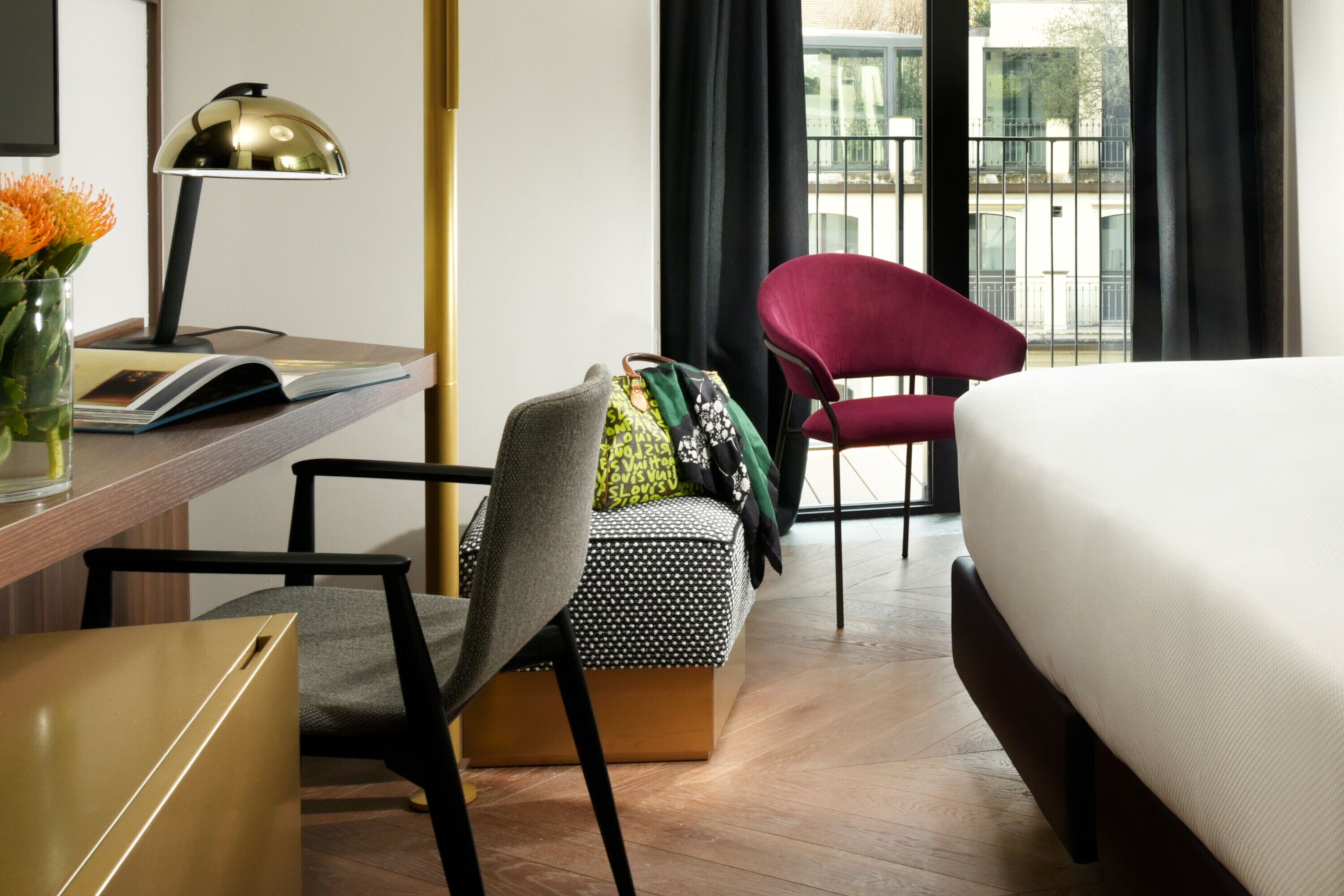 Une chambre d’hôtel avec un lit, un bureau avec une lampe, une chaise, une petite table et une fenêtre avec des rideaux.