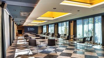 Un salón de conferencias con suelo a cuadros, filas de mesas y sillas, y amplias ventanas con cortinas.