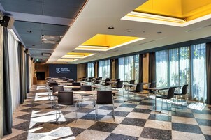Una sala conferenze con pavimento a scacchi, file di tavoli e sedie, e ampie finestre con tende.
