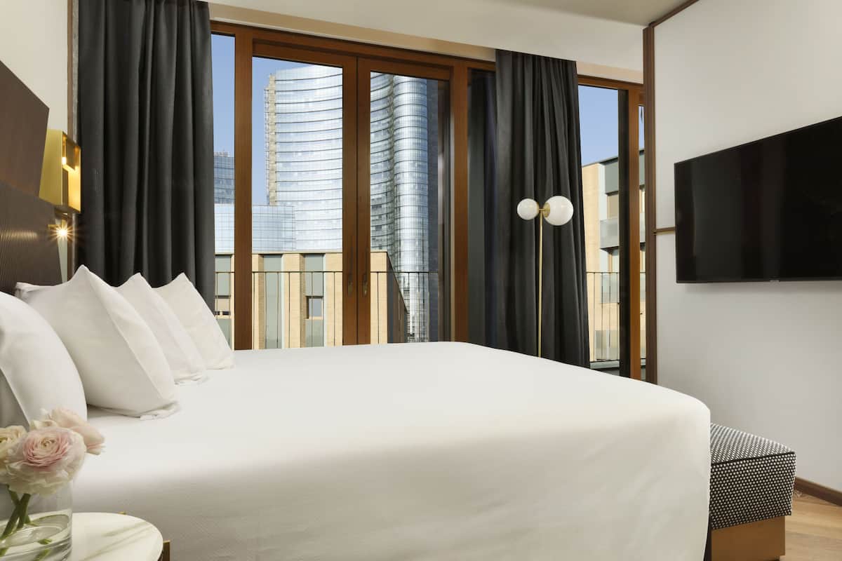 Une chambre d’hôtel moderne dotée d’un grand lit, d’un téléviseur à écran plat et offrant une vue sur le paysage urbain grâce à de grandes fenêtres du sol au plafond.