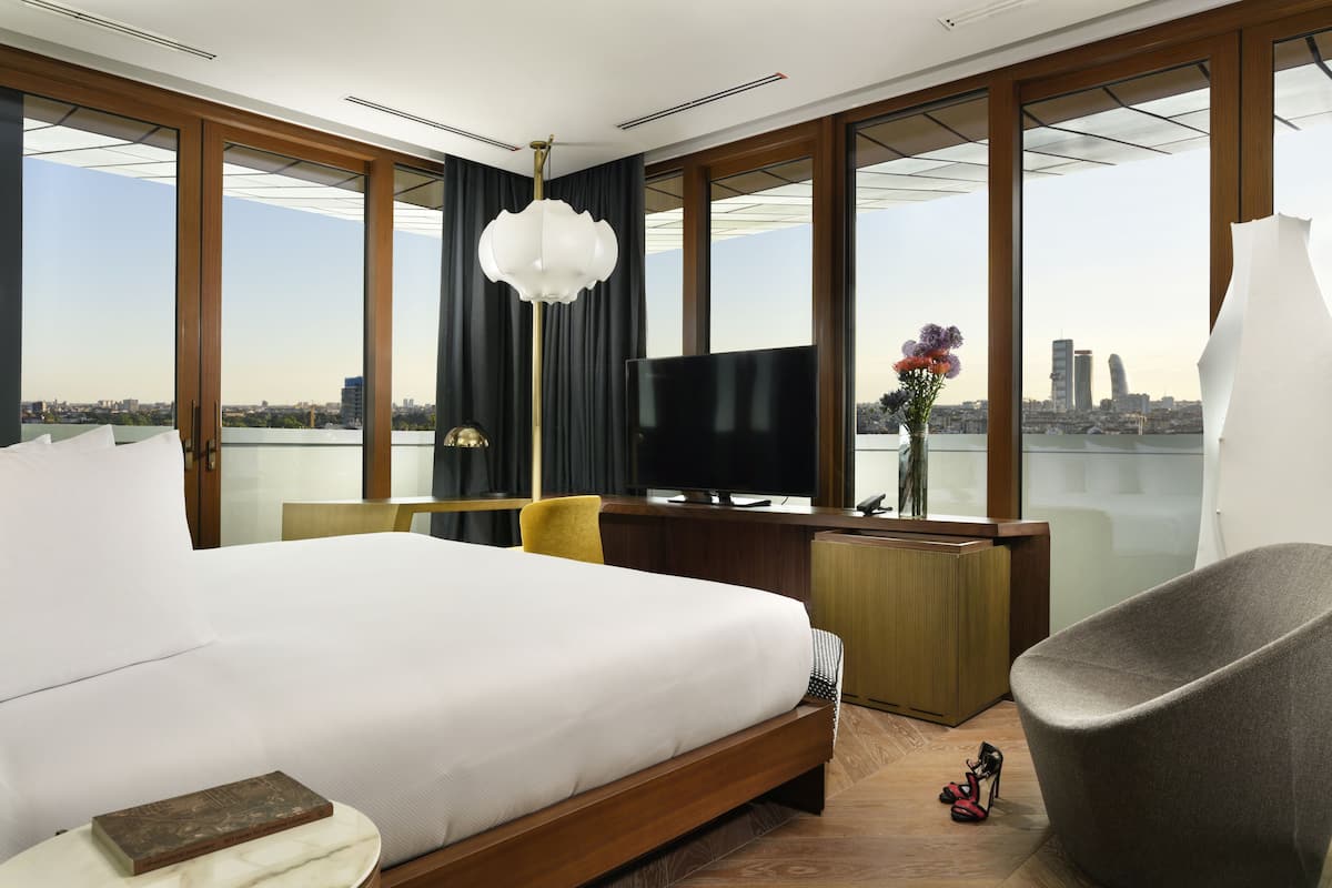 Une chambre d’hôtel moderne dotée d’un grand lit, d’un bureau avec un téléviseur à écran plat, d’une chaise et offrant une vue sur le paysage urbain.