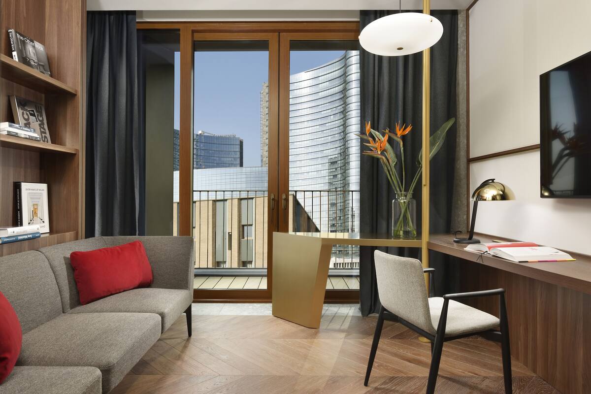 Un salon moderne avec un canapé, un bureau et une chaise. De grandes fenêtres offrent une vue sur le paysage urbain.