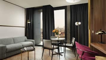 Un salon moderne avec un canapé gris, une petite table ronde et une table basse. On y trouve deux chaises, une grise et une rose, ainsi qu’un vase avec des fleurs sur la table. La pièce est dotée de grandes fenêtres avec des rideaux noirs, et offre une vue sur le paysage urbain. Le sol est en bois.
