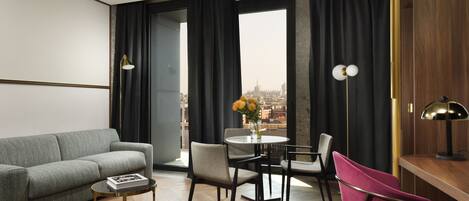 A modern living room with a grey sofa, a small round table, and a coffee table. There are two chairs, one grey and one pink, and a vase with flowers on the table. The room has large windows with black curtains, a view of a cityscape, and a wooden floor.